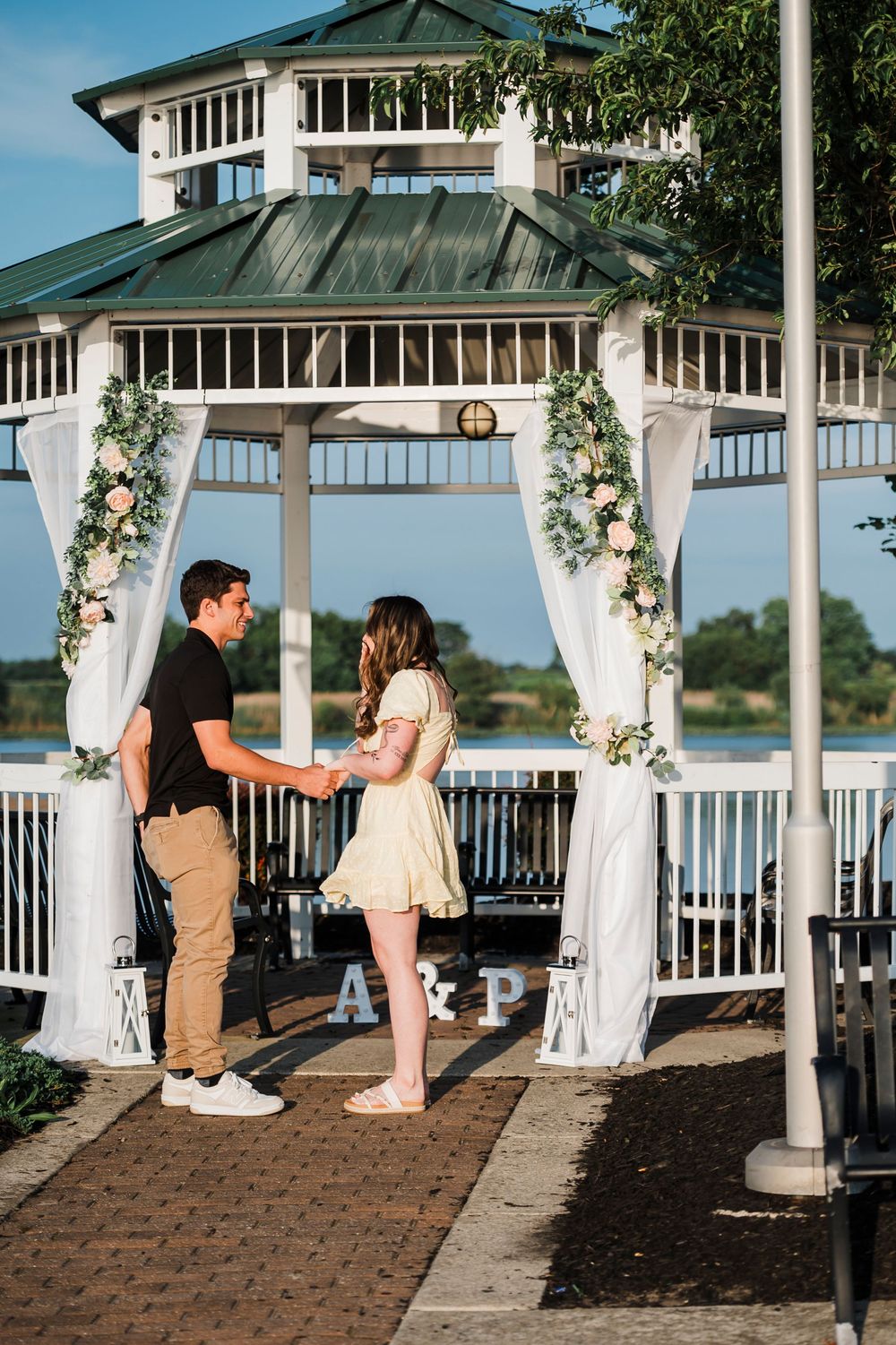 A couple shares a romantic moment in a white gazebo decorated with floral arrangements by the waterfront.