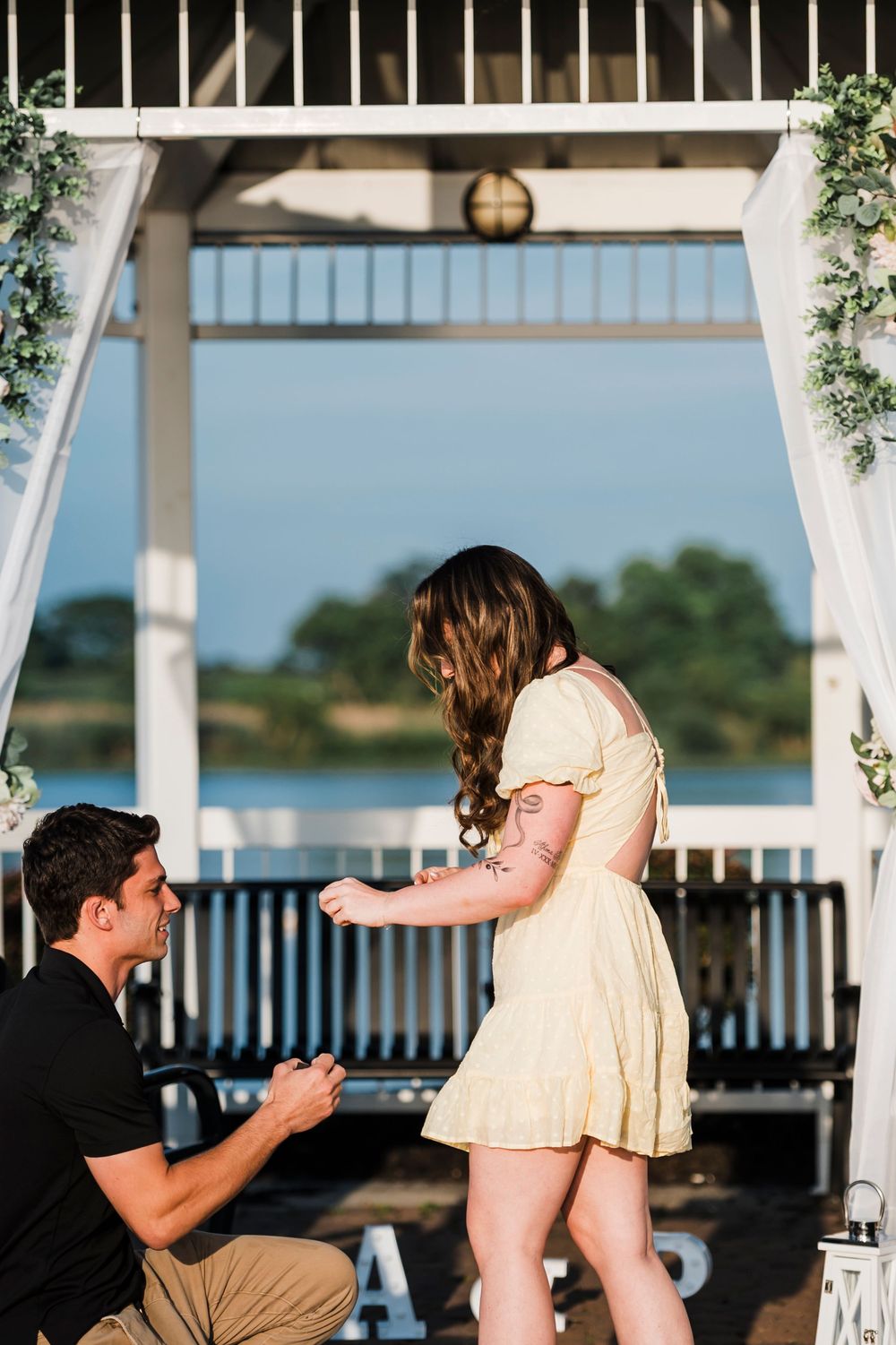 A romantic marriage proposal scene unfolds under a white gazebo by the waterfront at sunset.