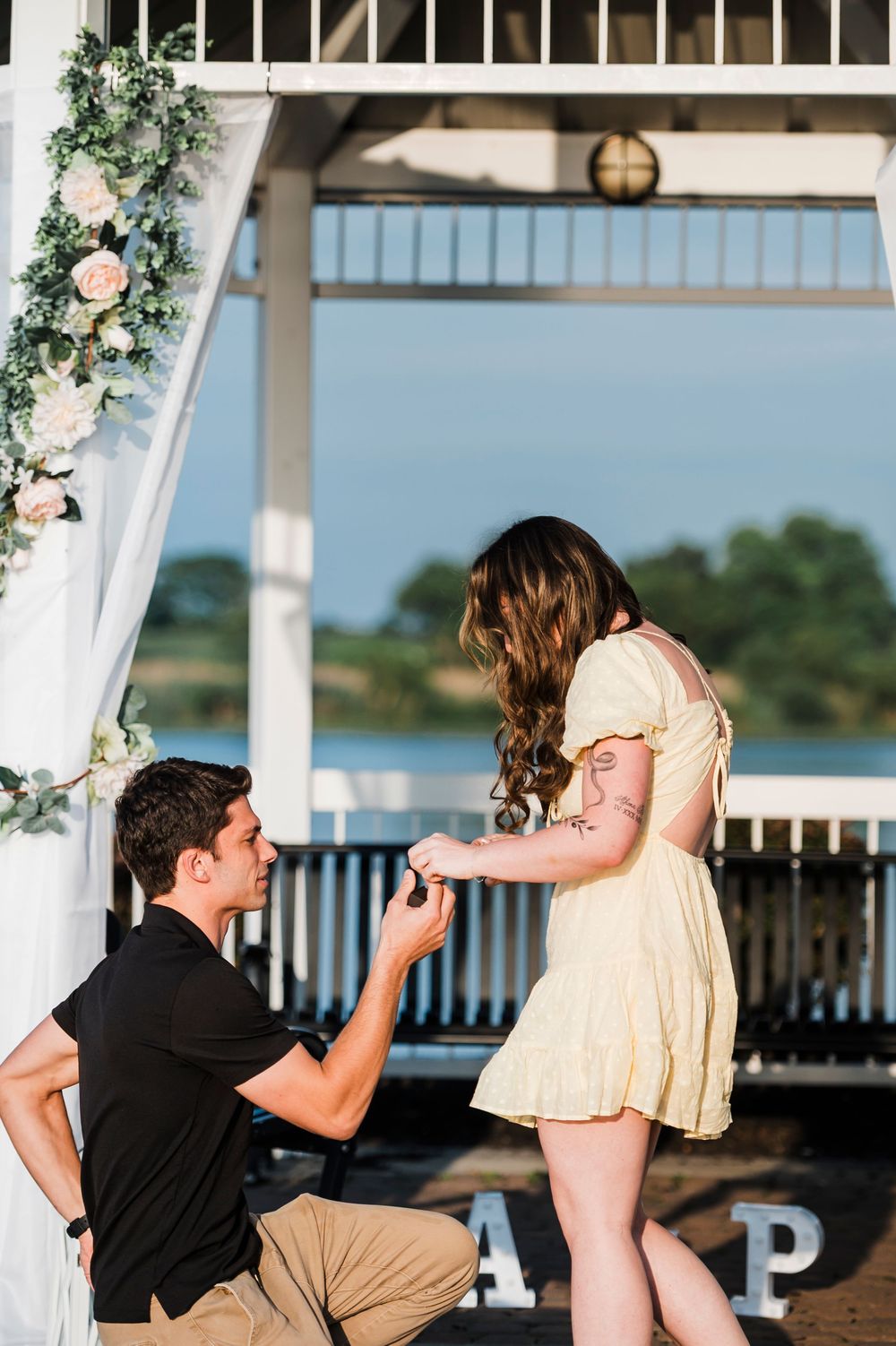 A joyful engagement moment captured in sequence at a lakeside gazebo adorned with white drapes and pink flowers.