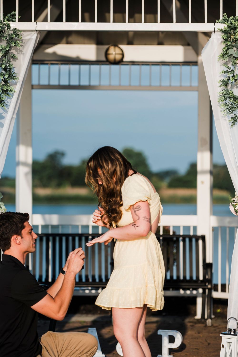 A romantic proposal moment captured at a waterfront gazebo with a yellow dress and kneeling pose.