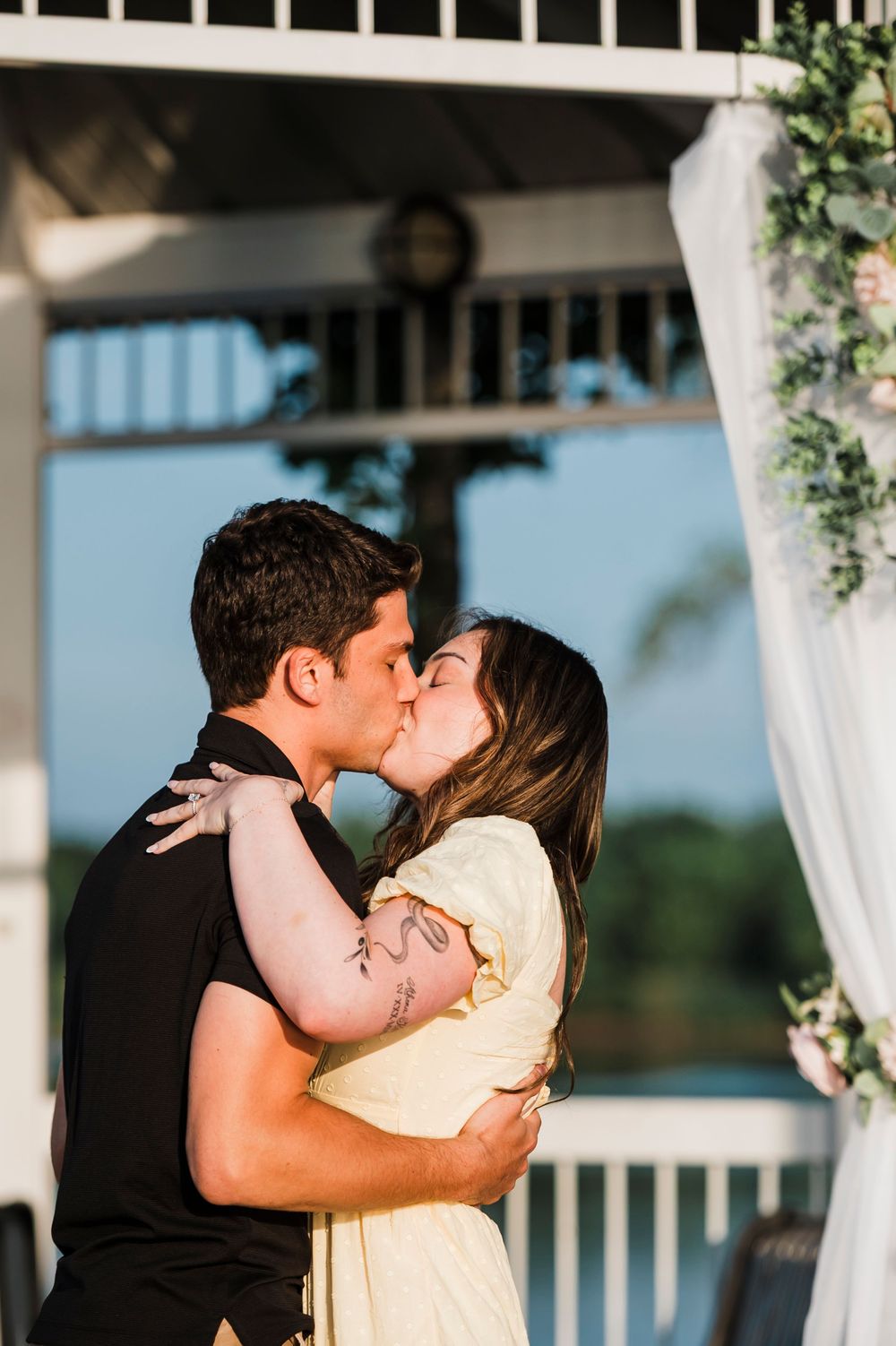 A romantic moment captured between a couple sharing a kiss on a waterfront gazebo during sunset.