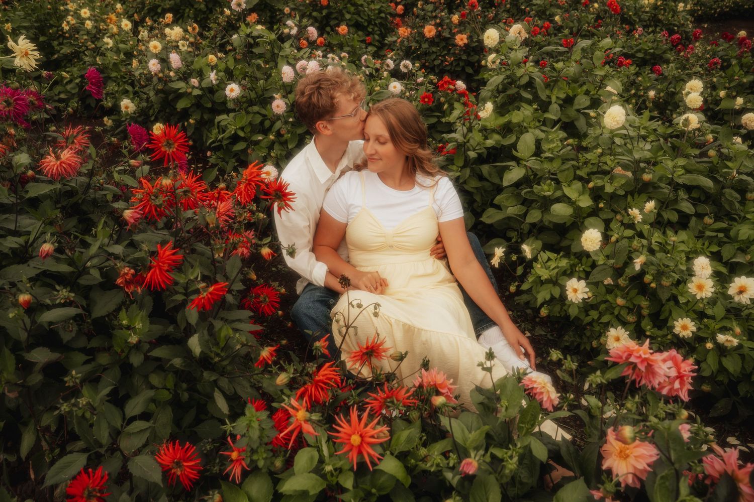 Romantic couple sitting together surrounded by vibrant red and pink dahlia flowers in a garden.