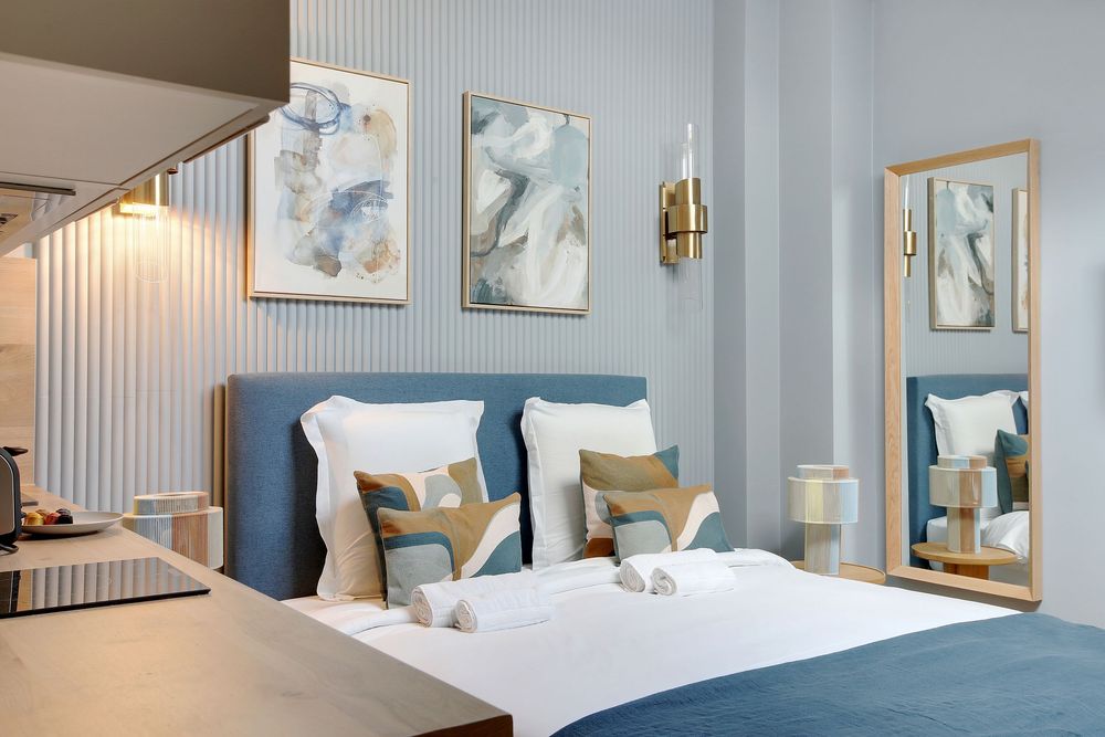 Stylish hotel room featuring a blue tufted headboard, patterned cushions, gold sconce, and wall mirror.