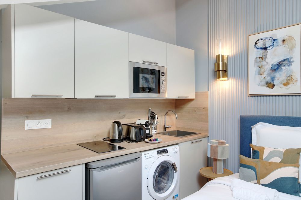 Modern studio kitchenette with white cabinets, built-in microwave, washing machine, and blue upholstered bed nearby.
