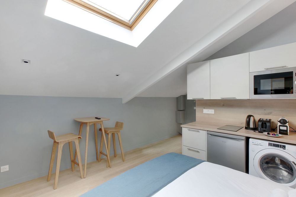 Bright studio apartment with skylight, white kitchen cabinets, washing machine, and small wooden dining table with bar stools.