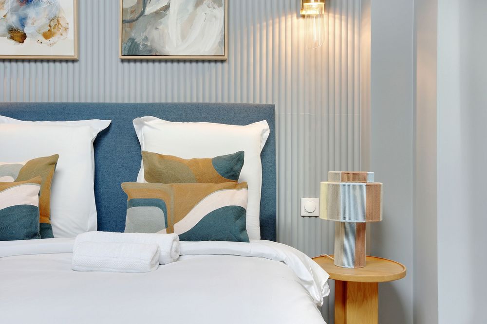Close-up of luxury bed with blue headboard, decorative pillows, white towels, and wooden bedside lamp.