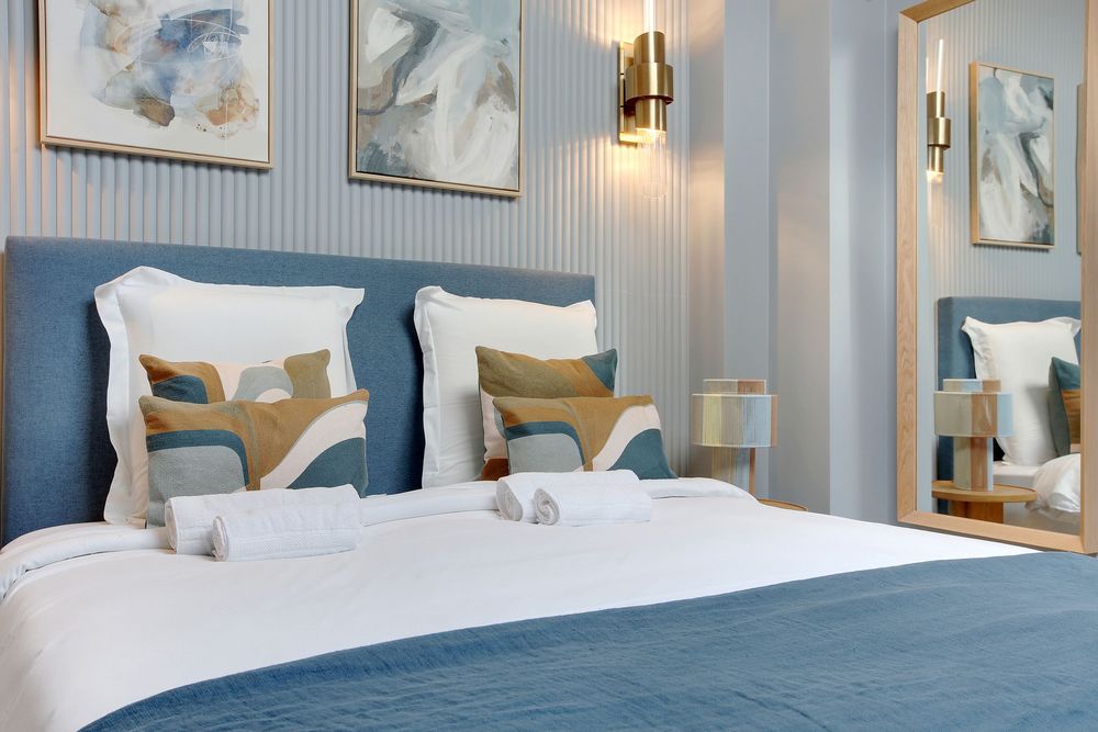 Elegant double bed with blue velvet headboard, patterned cushions, white linens, and gold wall lighting.