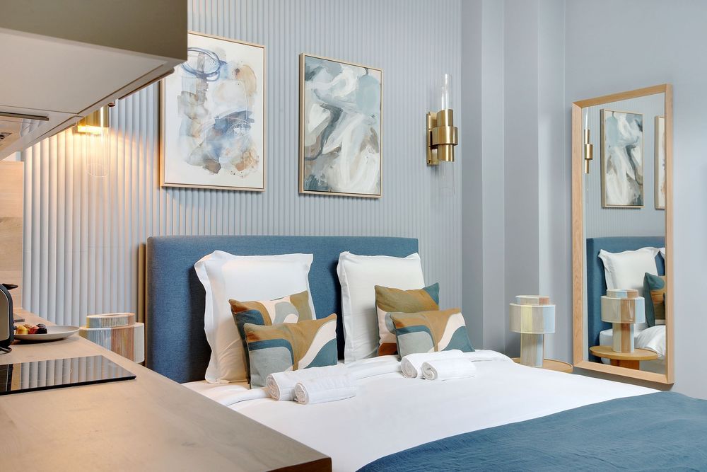 Luxury hotel bedroom with blue headboard, colorful decorative pillows, gold lighting, and framed artwork.