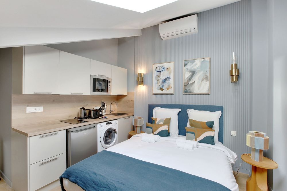 Compact studio apartment with blue bed, kitchenette, washing machine, and modern minimalist decor.