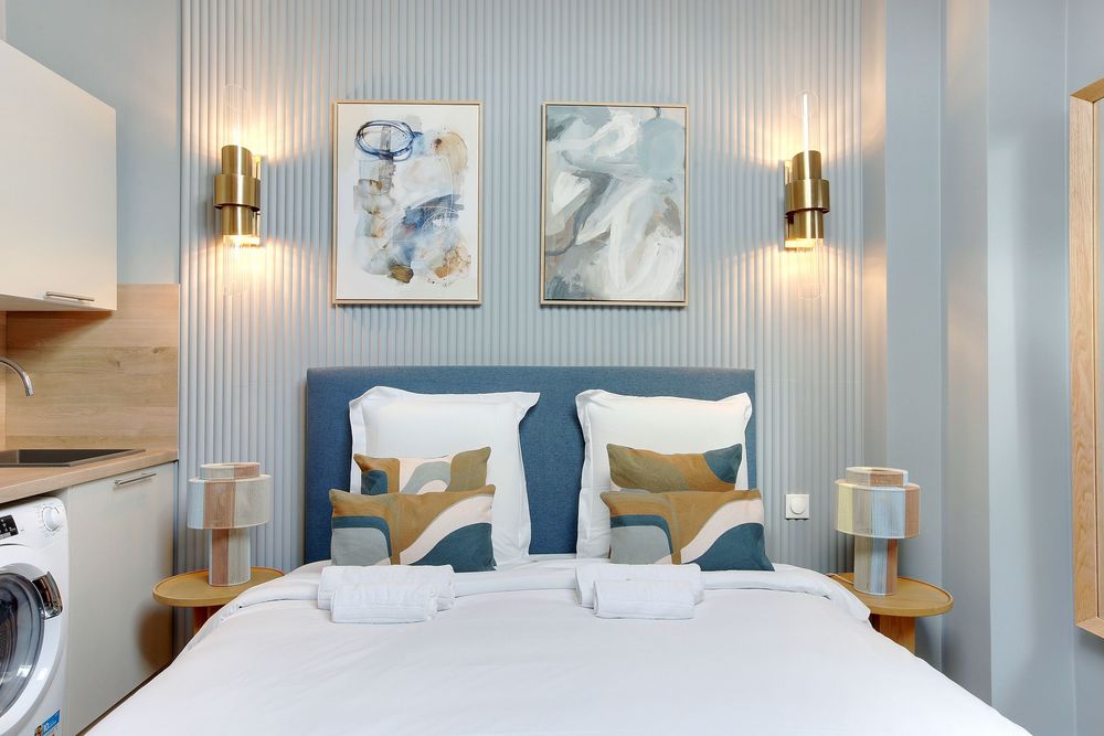 Stylish studio bedroom with blue tufted headboard, decorative pillows, gold wall sconces, and abstract artwork above bed.