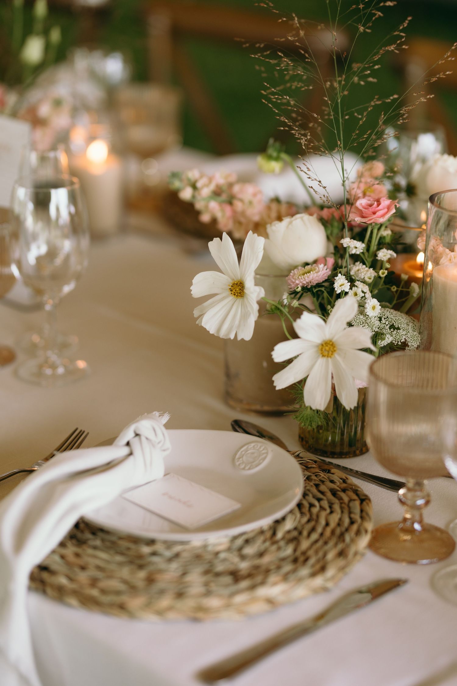 Elegant table setting with white daisies in vase, woven placemat, white plate and wine glass on linen tablecloth.