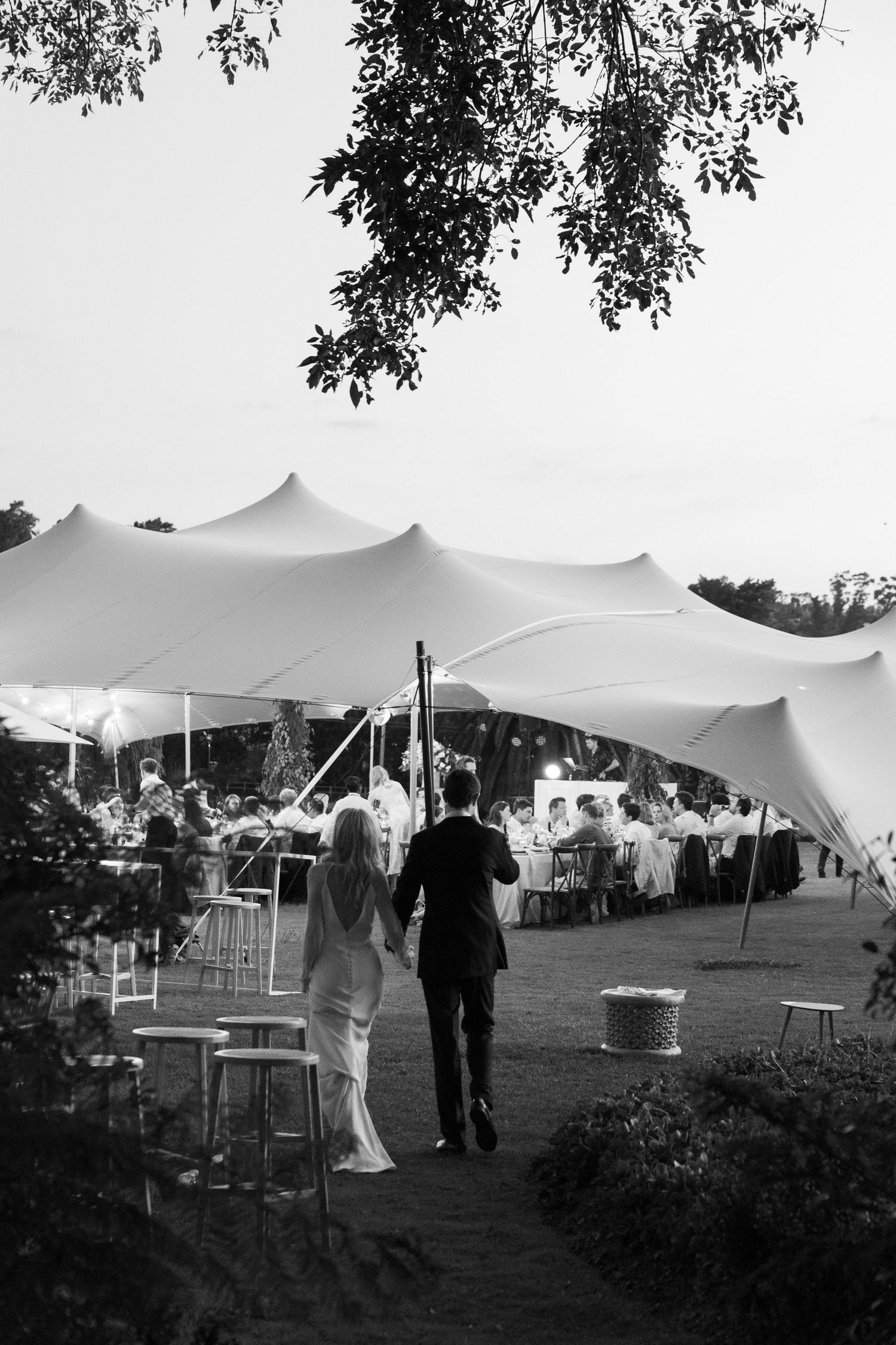 A couple walks toward an elegant outdoor wedding reception under flowing white tent canopies in black and white photography.