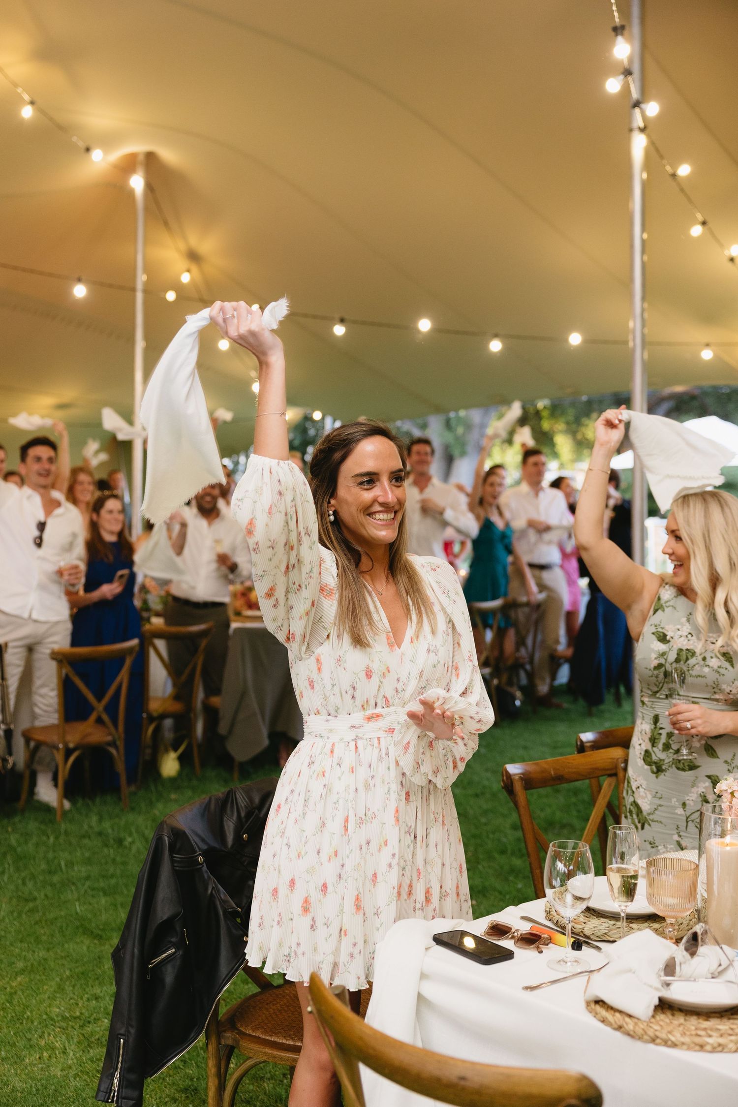 Guests celebrate at an outdoor reception under string lights with a white tent and wooden chairs on artificial grass.