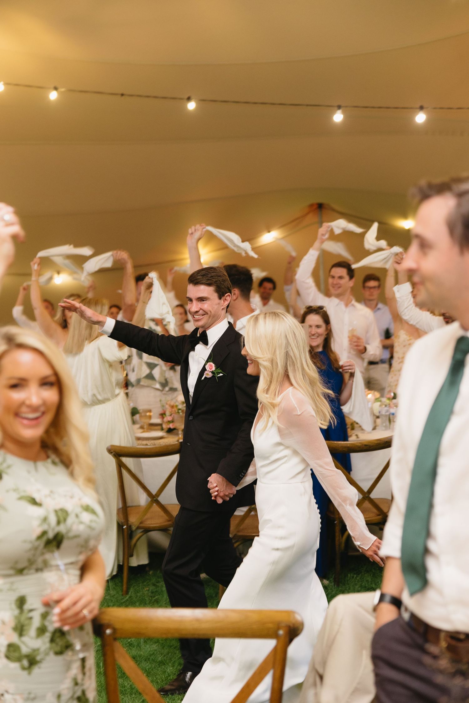 Wedding celebration under string lights with guests dancing and celebrating on a grassy outdoor venue.