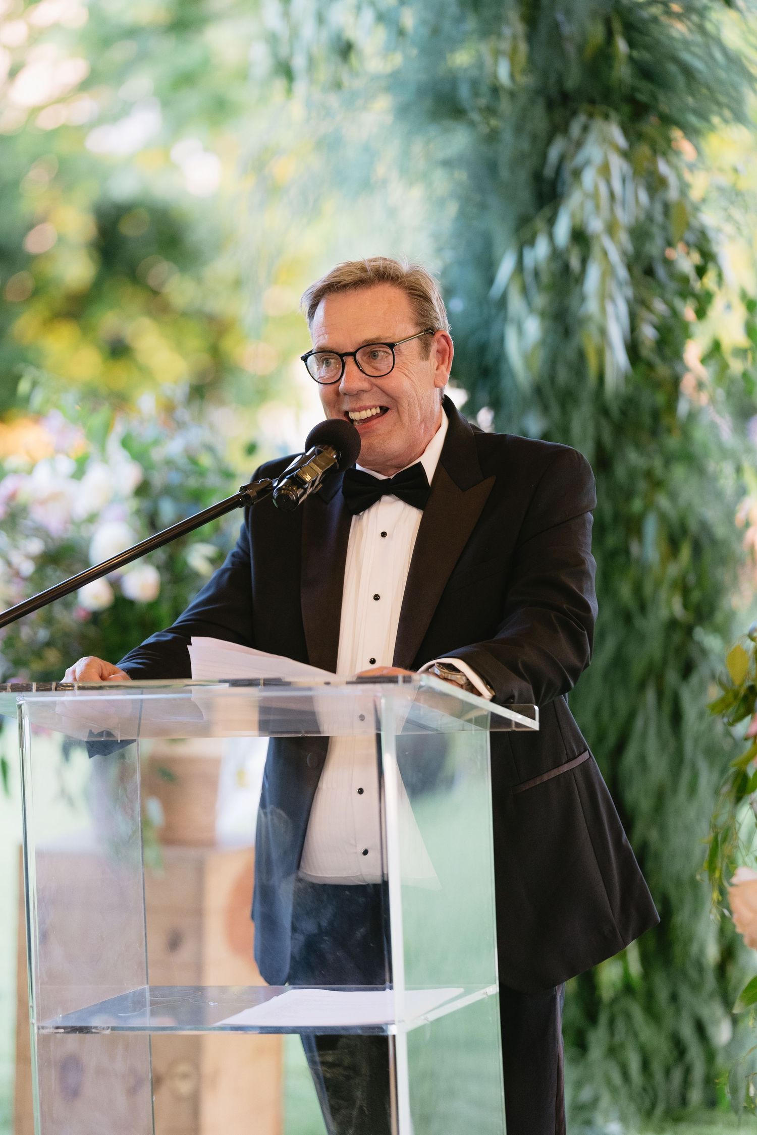A speaker in a black tuxedo and glasses stands at a clear acrylic podium during a formal event.
