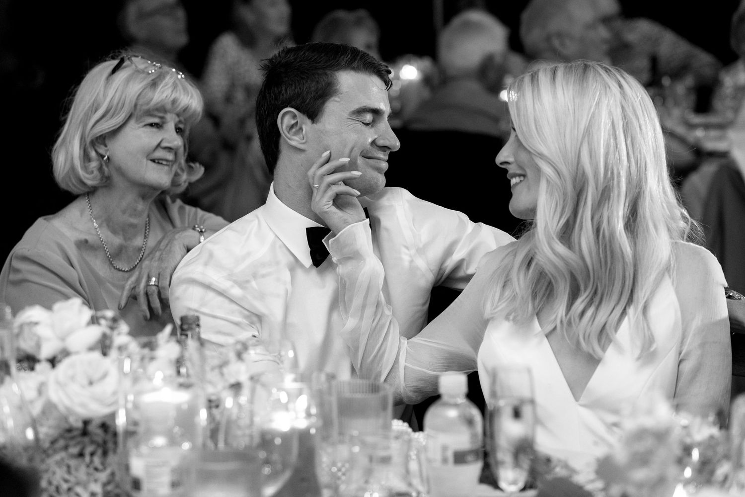 Three people share a candid moment at an elegant dinner table with glowing candles in black and white.