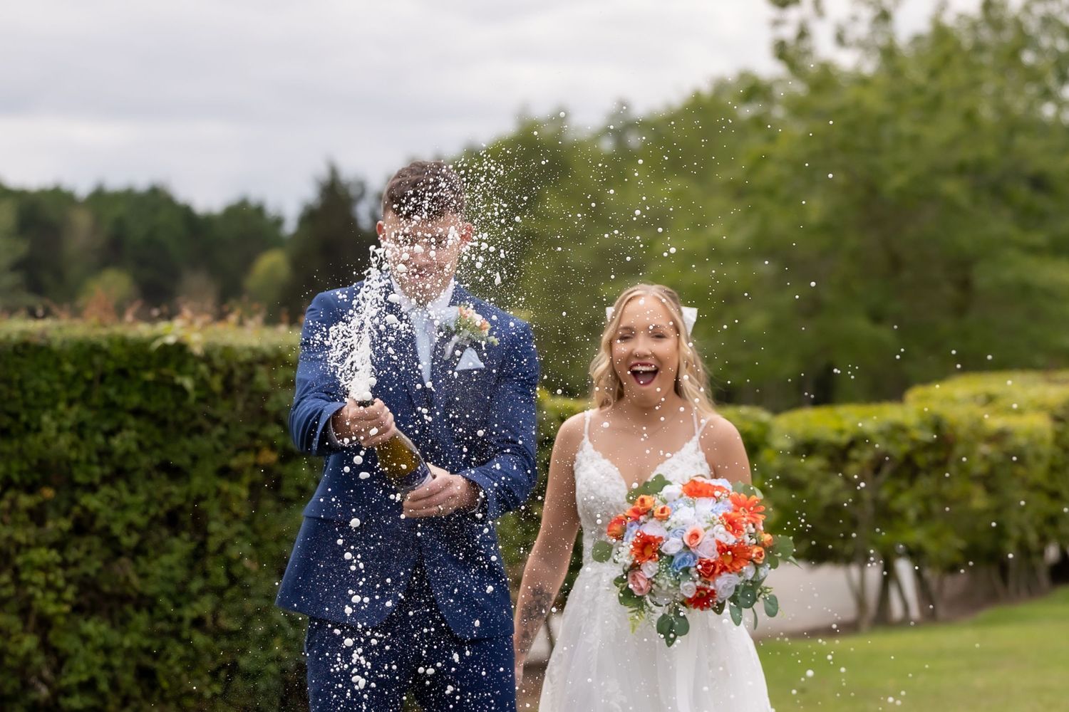 A wedding couple walks through a garden path while spraying champagne from a bottle.