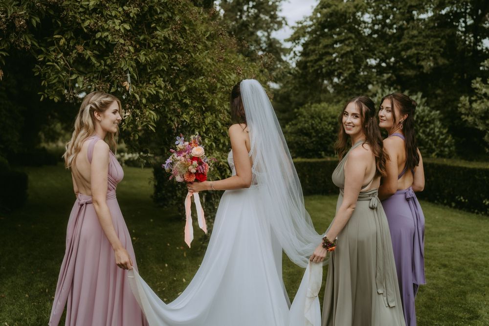 Bridal party in lavender and pink dresses poses together on grassy lawn.