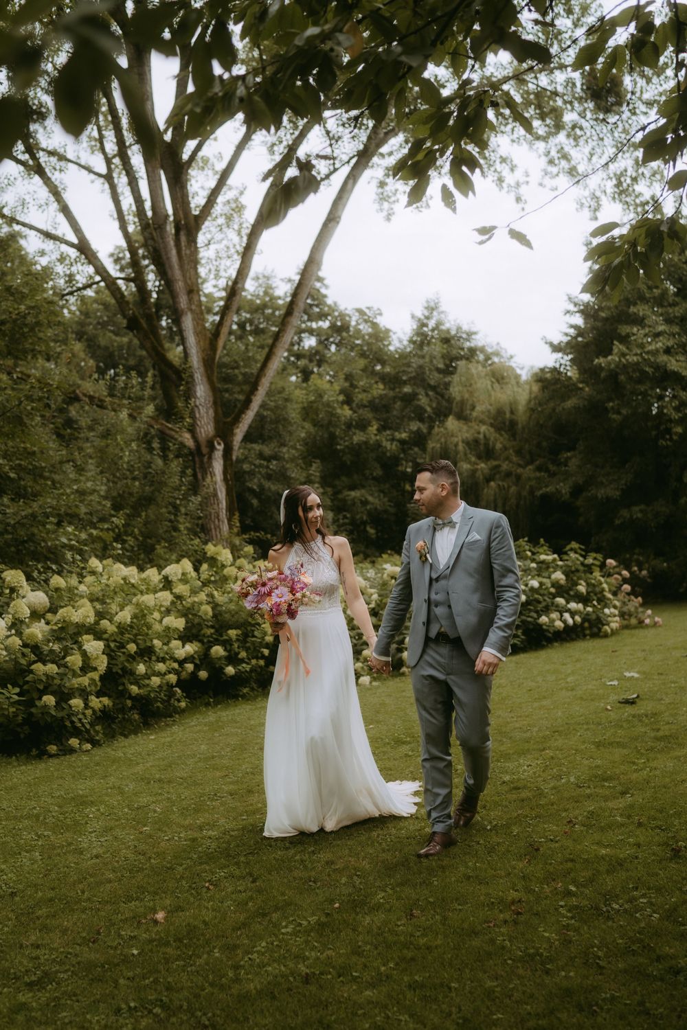 Newlyweds walk hand in hand through lush garden landscape.