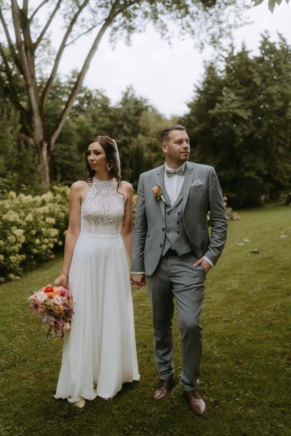 Bride in white dress and groom in gray suit pose in garden setting.