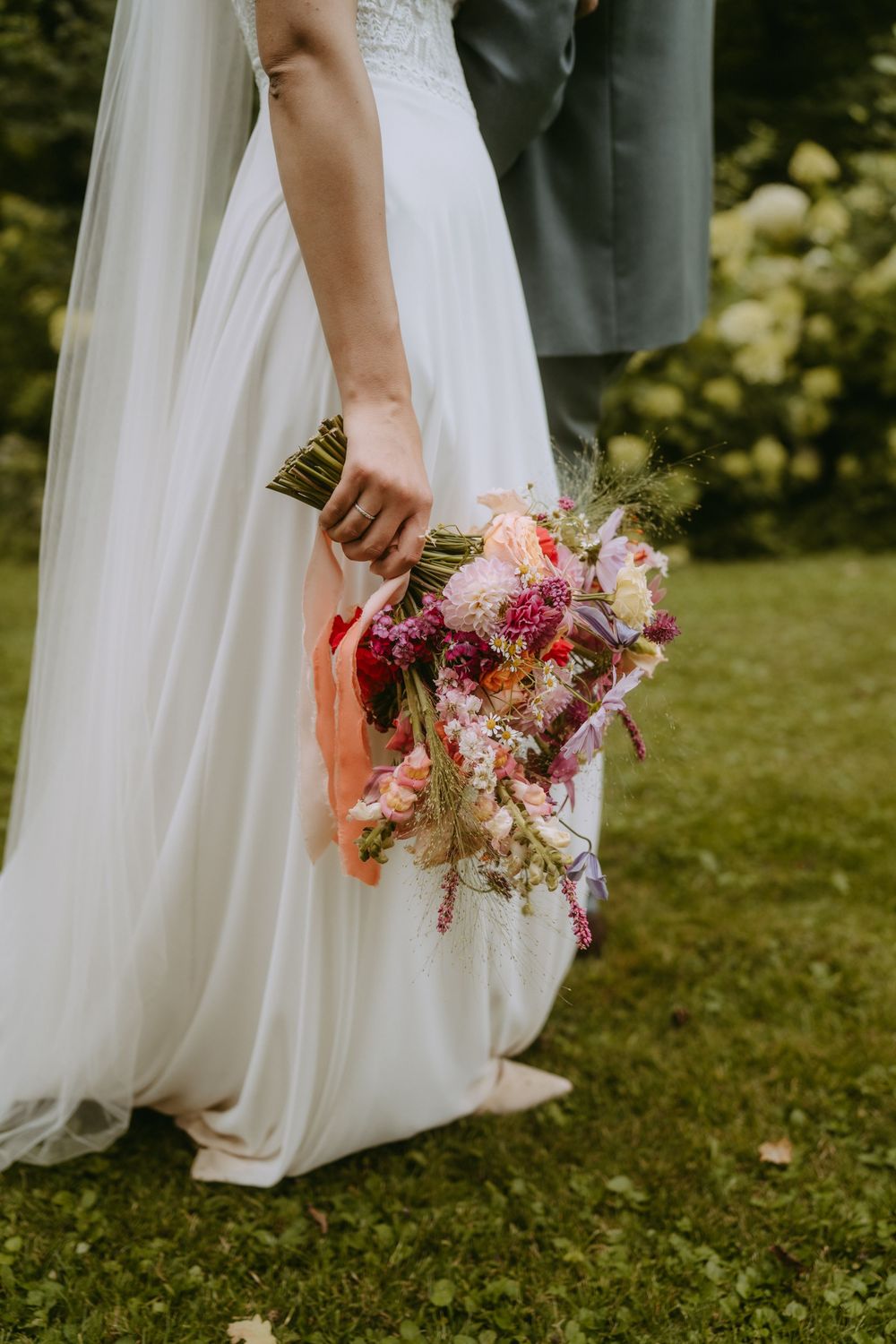 A colorful wedding bouquet with pink and peach flowers held against a flowing white dress in an outdoor garden setting.