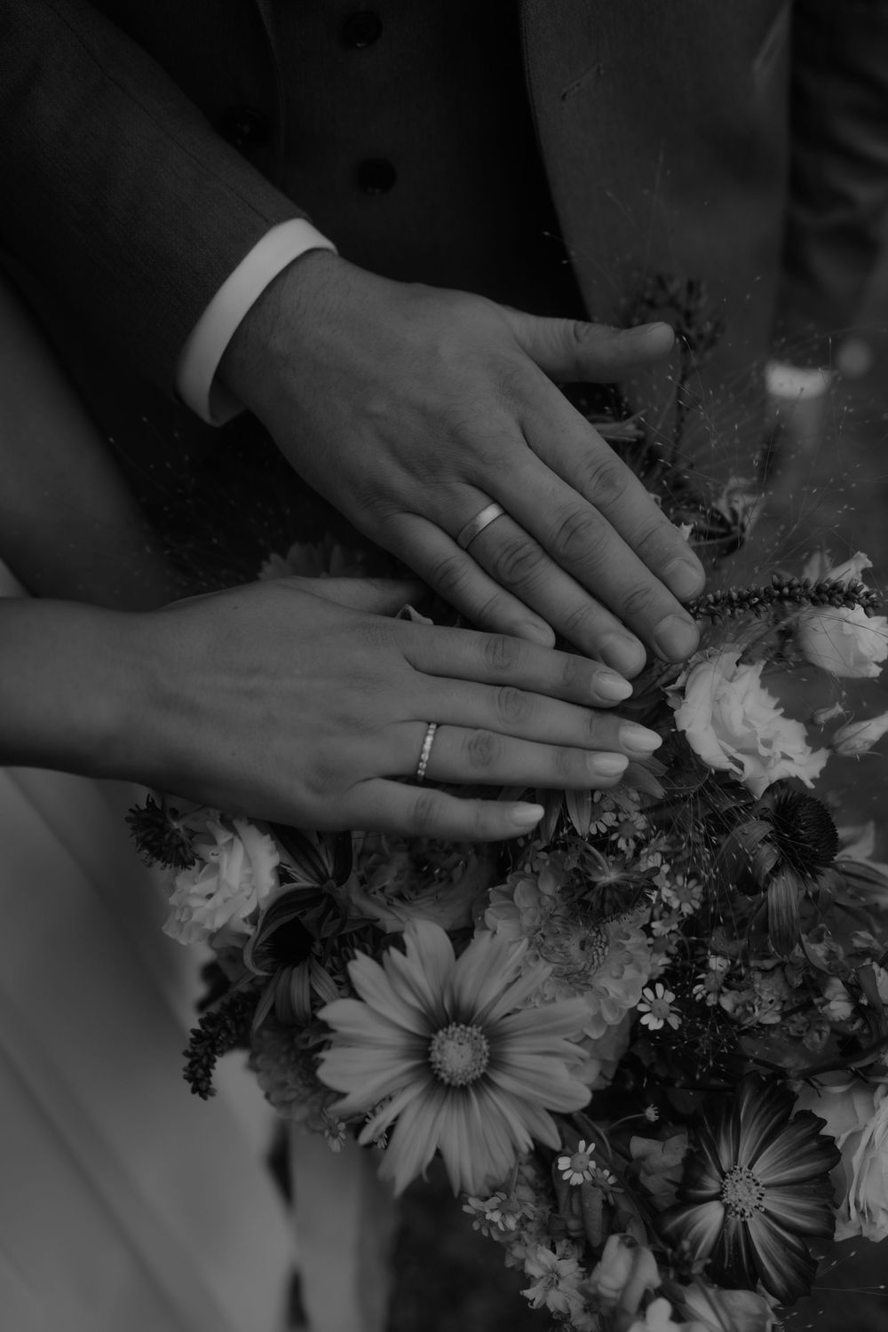 Black and white close-up of hands touching delicate flowers and wedding rings.