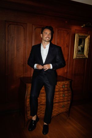 A person in a black suit and white shirt sits in a dimly lit room with wood-paneled walls.