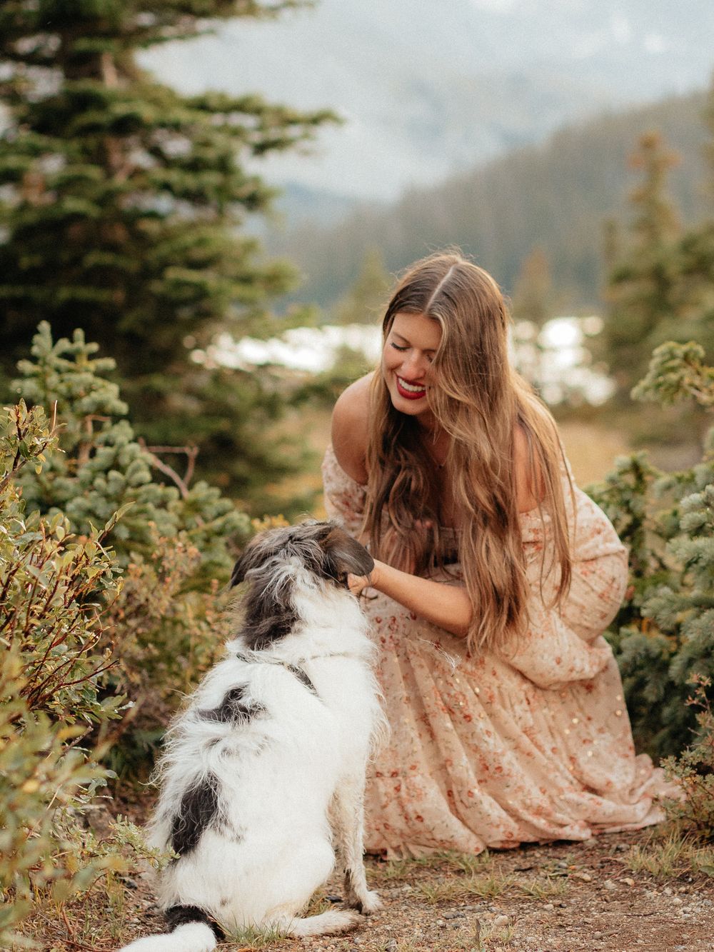 A person in a peach-colored dress shares a tender moment with their Australian Shepherd dog on a mountain trail.