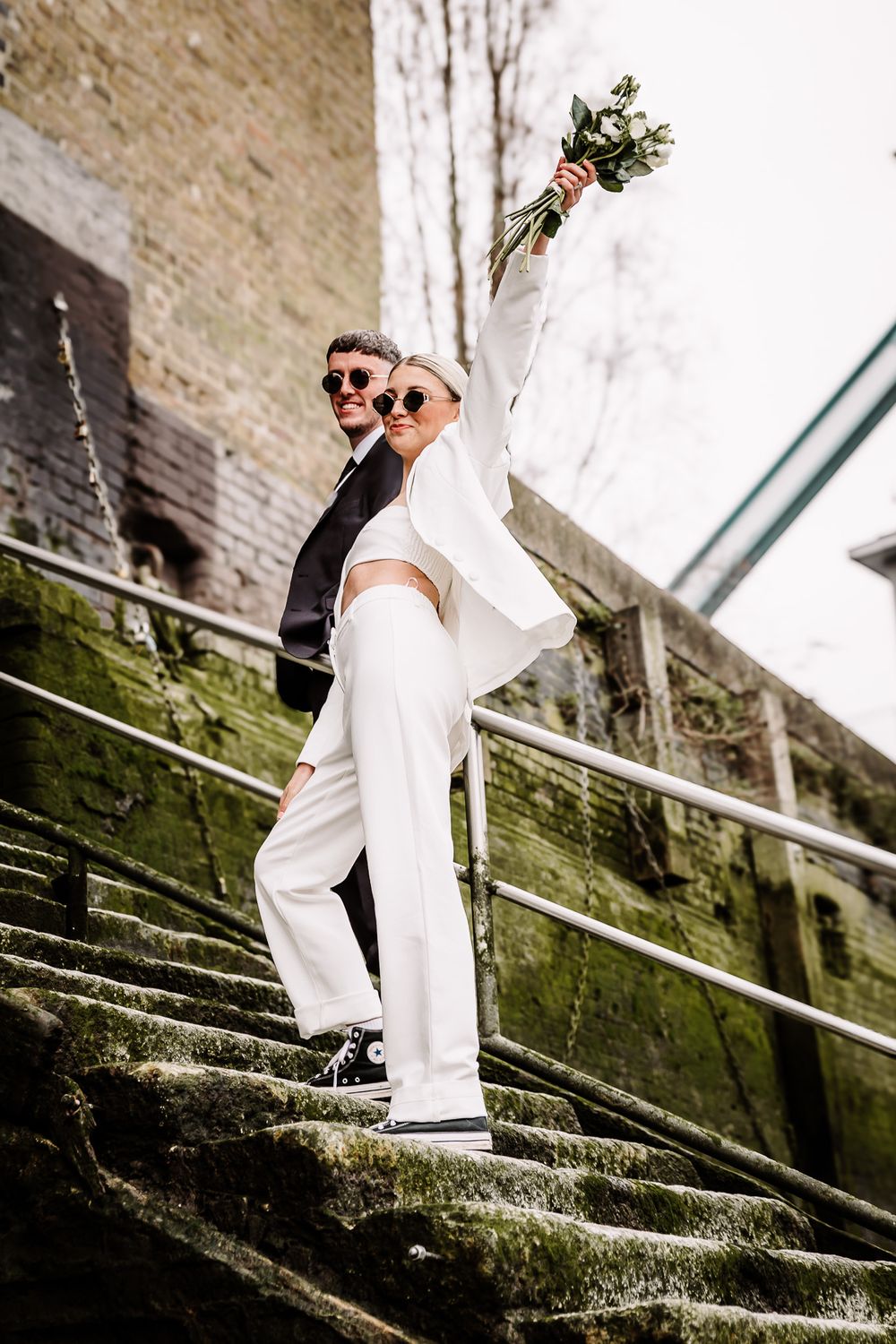 Couple in black and white attire walking up moss-covered stone steps in an urban setting.