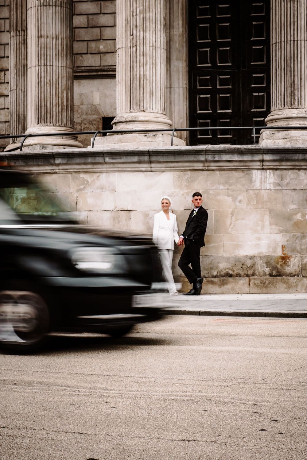 Wedding couple standing by classical columns as cars blur past in street.