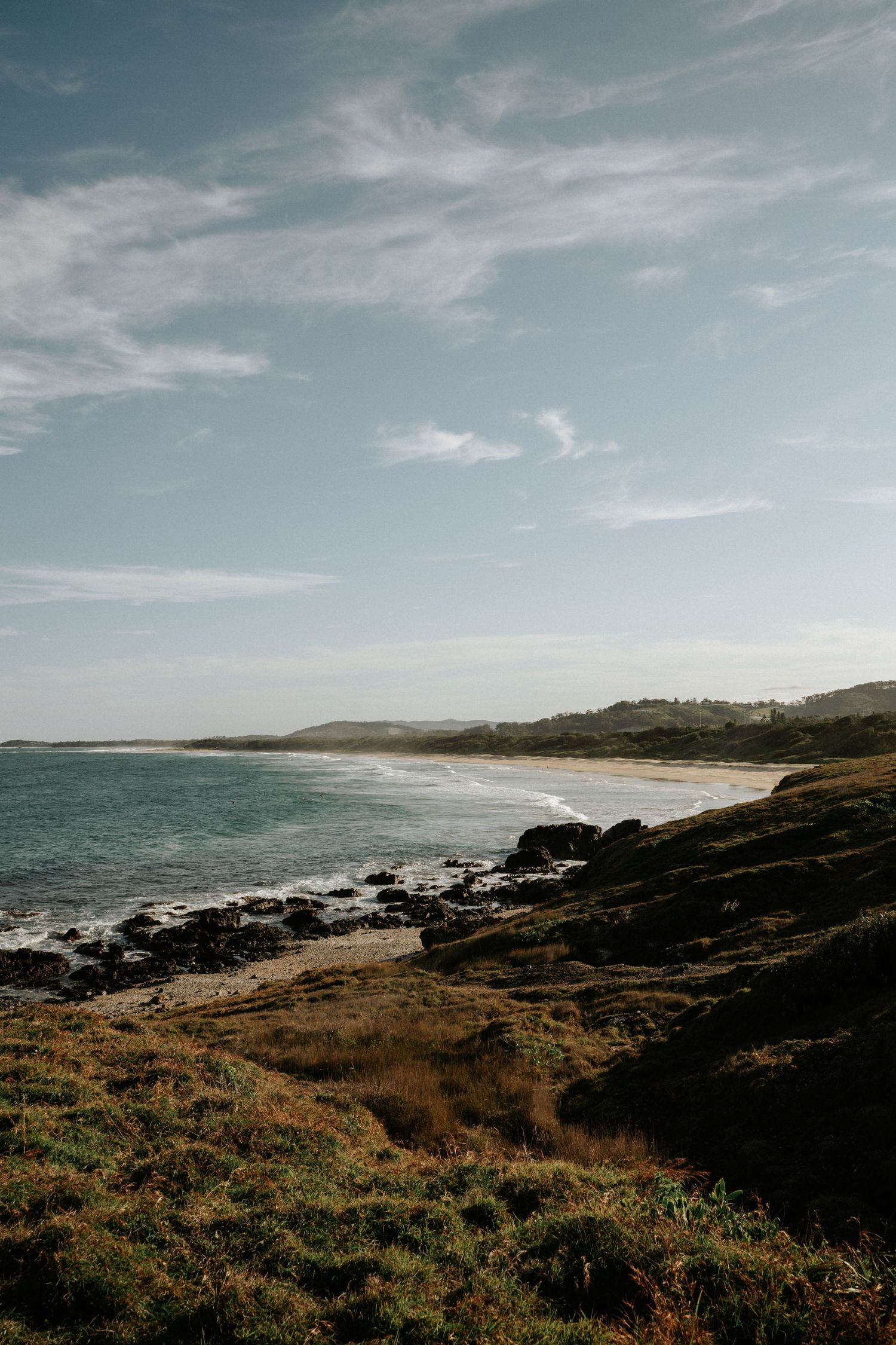 Rugged coastal landscape with rocky shoreline and grassy cliffs overlooking a serene ocean under wispy clouds.