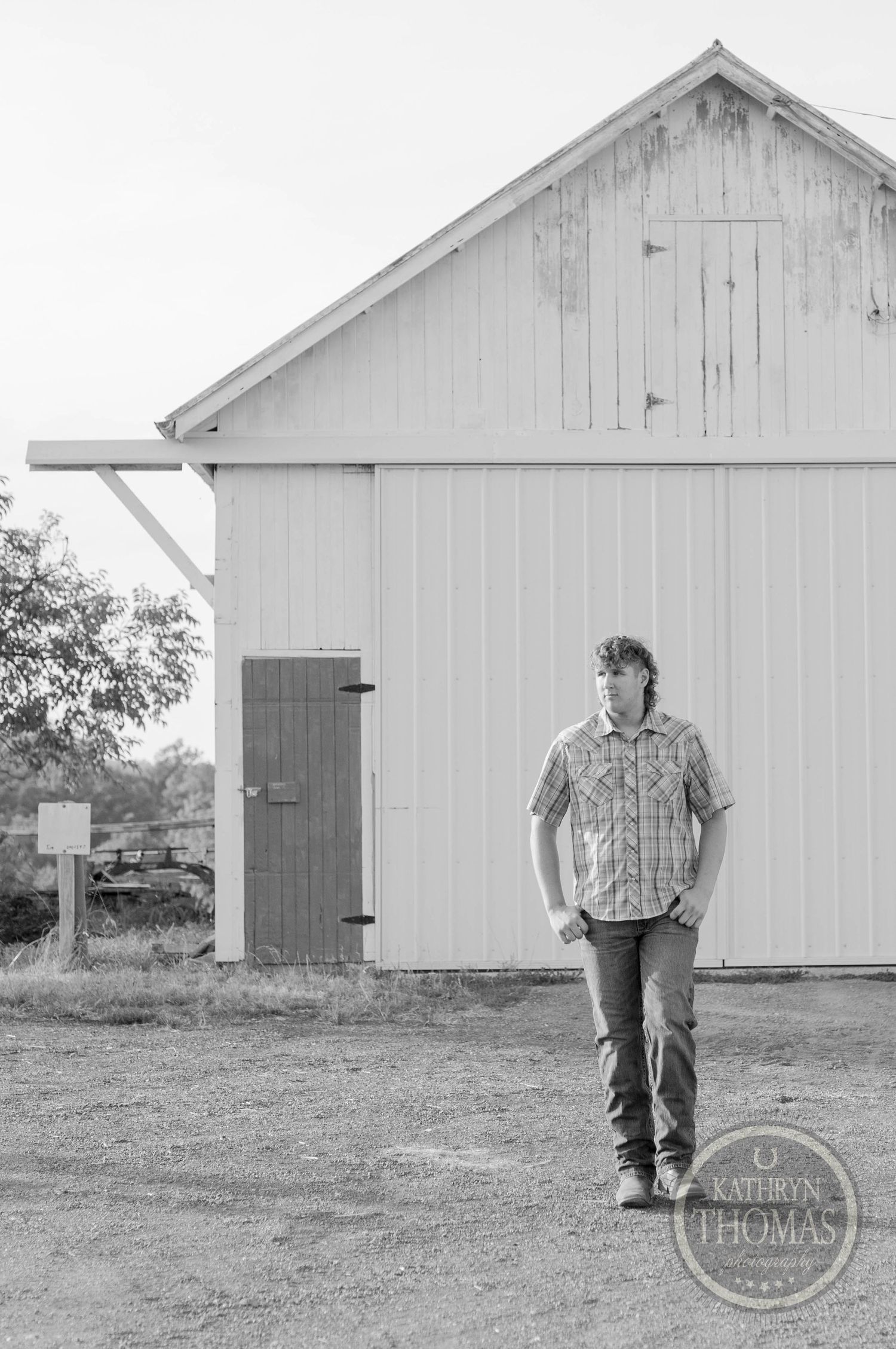 A black and white portrait standing beside a rustic white barn with vintage wooden doors.