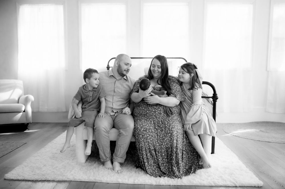A family of four sits together in a bright living room on a white rug for a black and white portrait session.