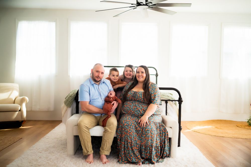 A family shares a tender moment while sitting together against a metal bed frame in a sunlit room.