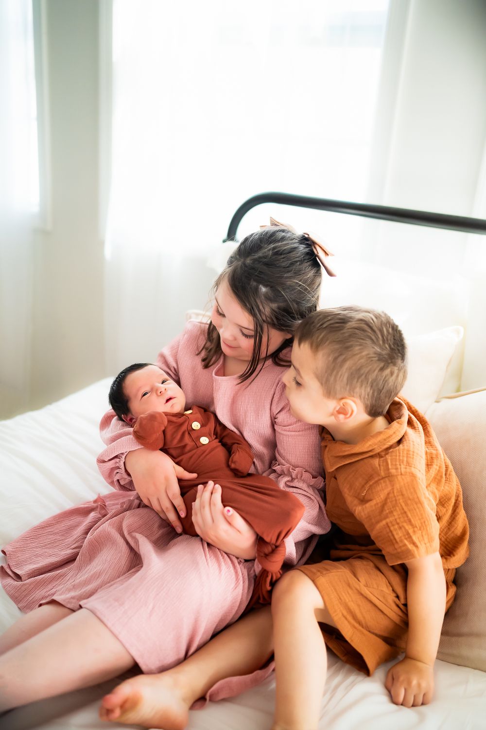 Two children and a baby in rust-colored and pink outfits share a tender moment while sitting together on a white couch.