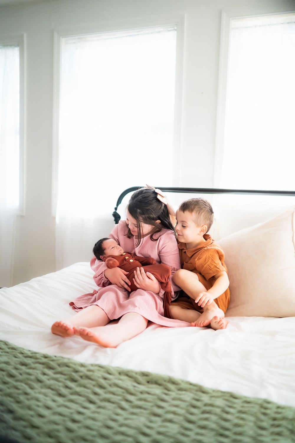 Three siblings snuggle together on a white bed with a green blanket in a bright sunlit room.