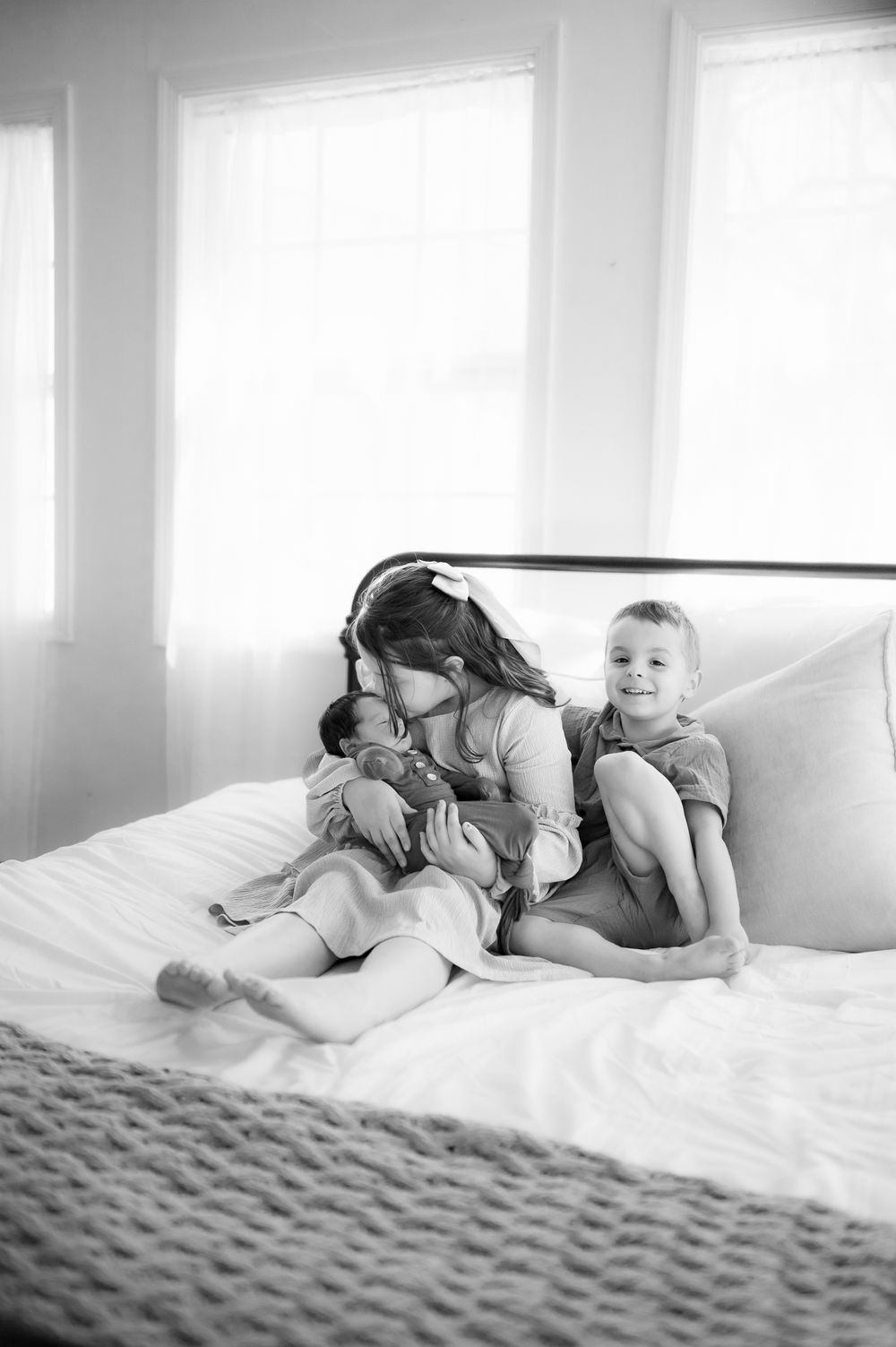 Two children relax together on a white bed near windows in a candid black and white photograph.