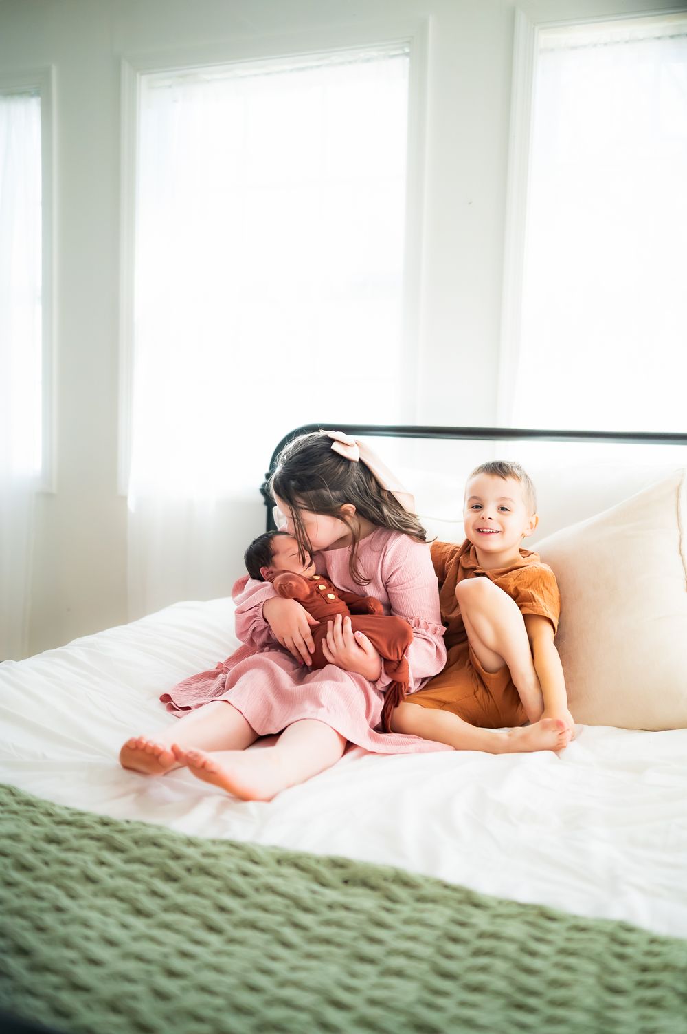 Children relaxing together on a white bed with green blanket in bright sunlit room wearing orange and pink outfits.