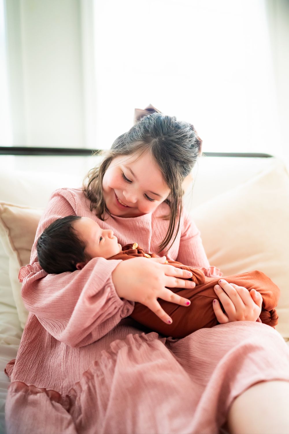 A tender moment captured as parent and infant share soft smiles while cuddling on couch in pink clothing.