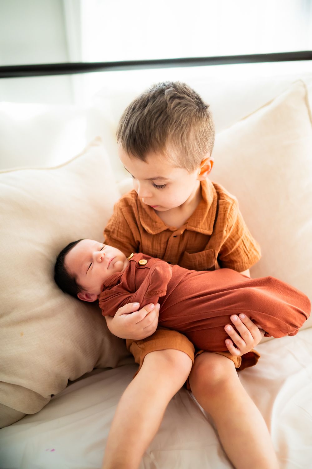 Two children wearing rust-colored outfits share a tender moment together on a white couch.