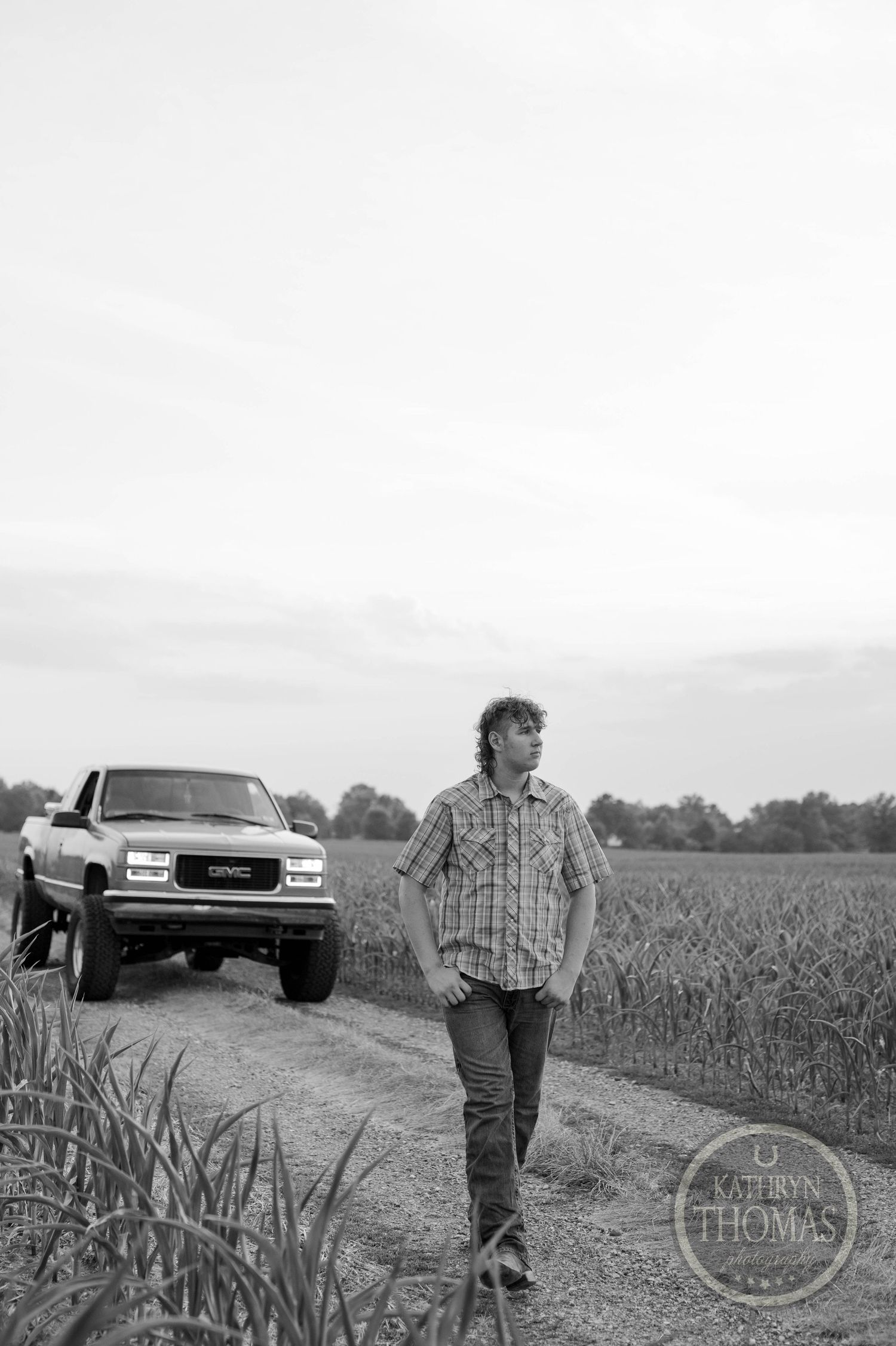 Black and white portrait of someone walking on rural path with lifted truck in background.
