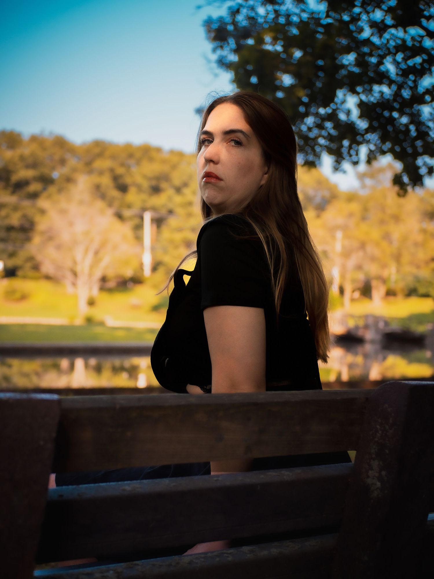 Someone with long dark hair sits thoughtfully on a park bench with autumn trees and a pond in the background.
