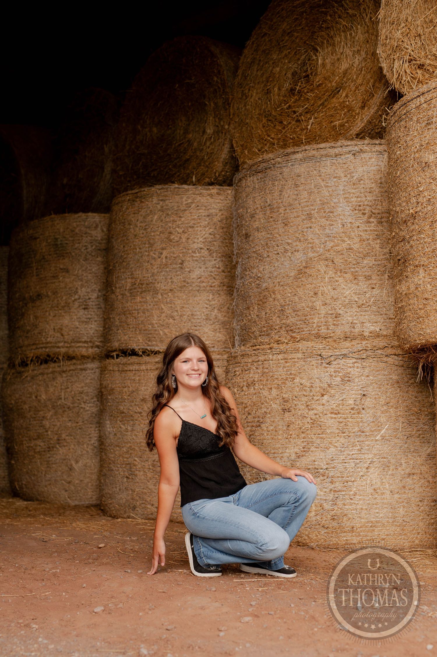 Rustic portrait sitting by large round hay bales in barn interior.