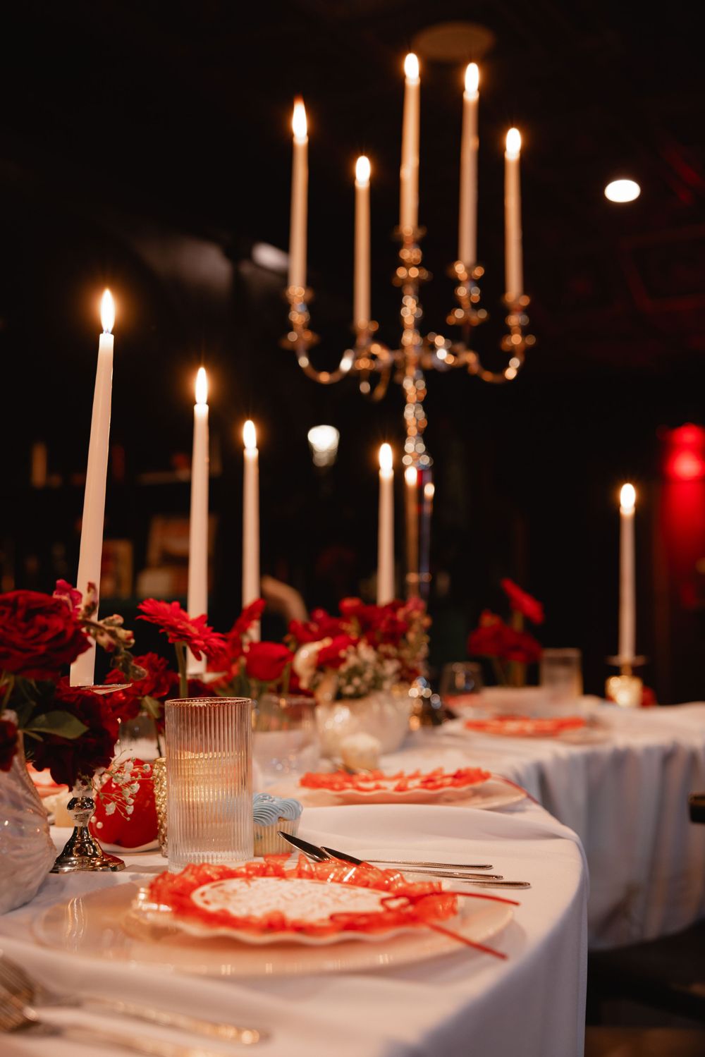 Elegant wedding reception table with white candles, red roses, and decorative red lace plates on white linen.
