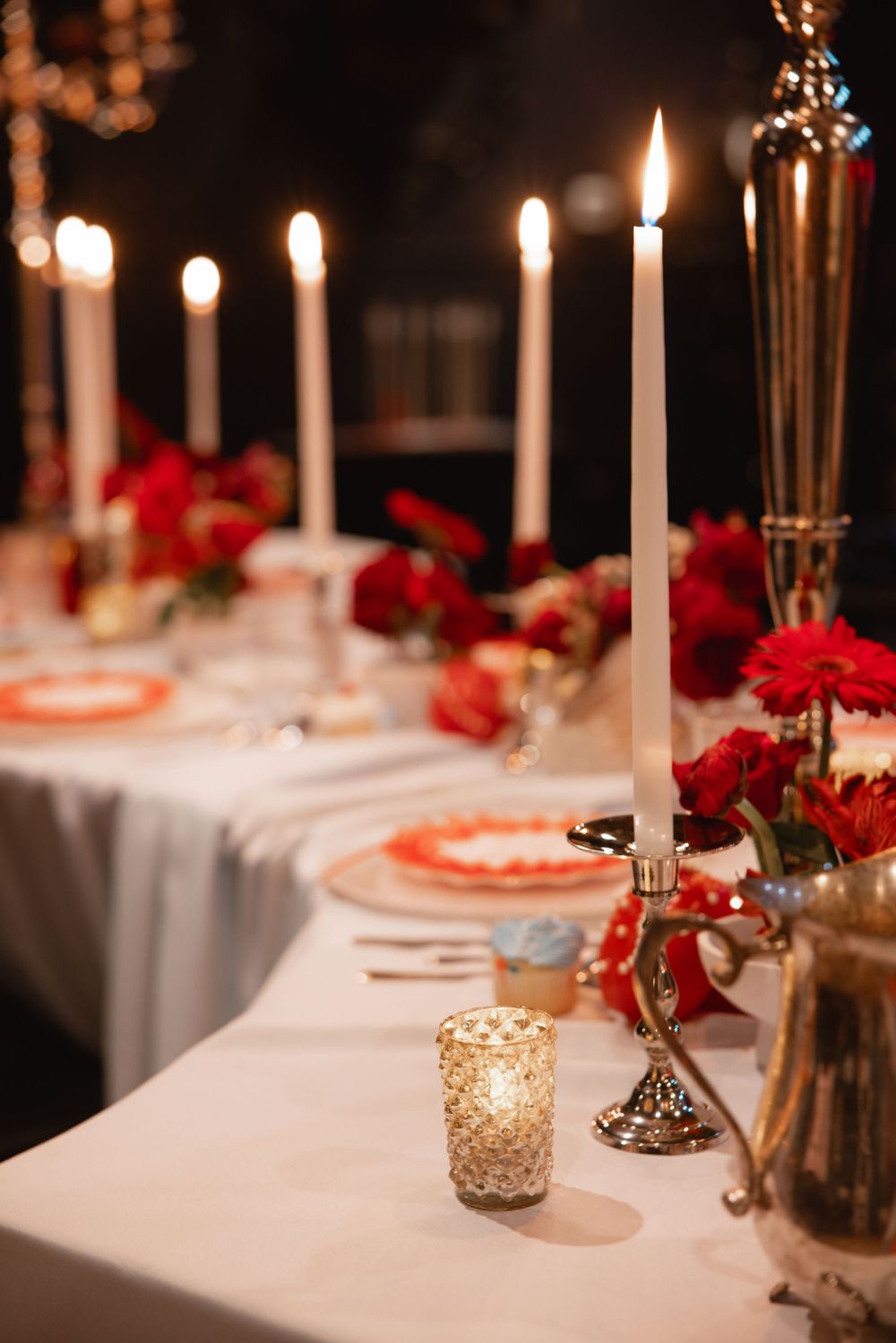 Candlelit wedding reception table with white taper candles, red floral arrangements, and gold votive holders.