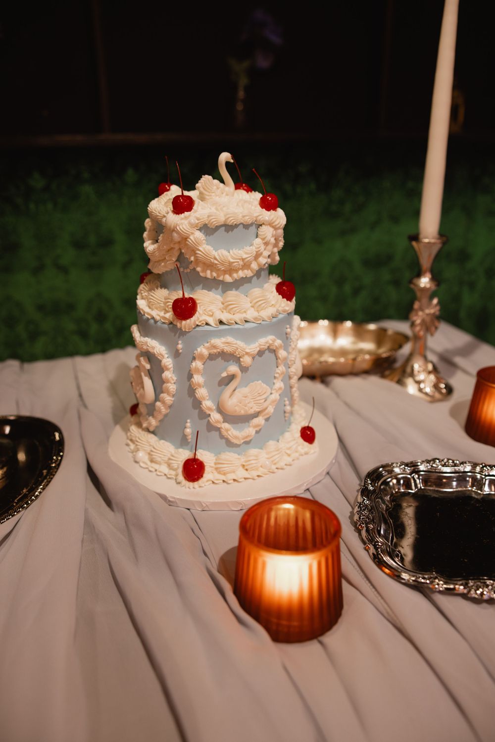 Two-tier light blue wedding cake with white swan and heart piping decorations and red cherry accents outdoors.