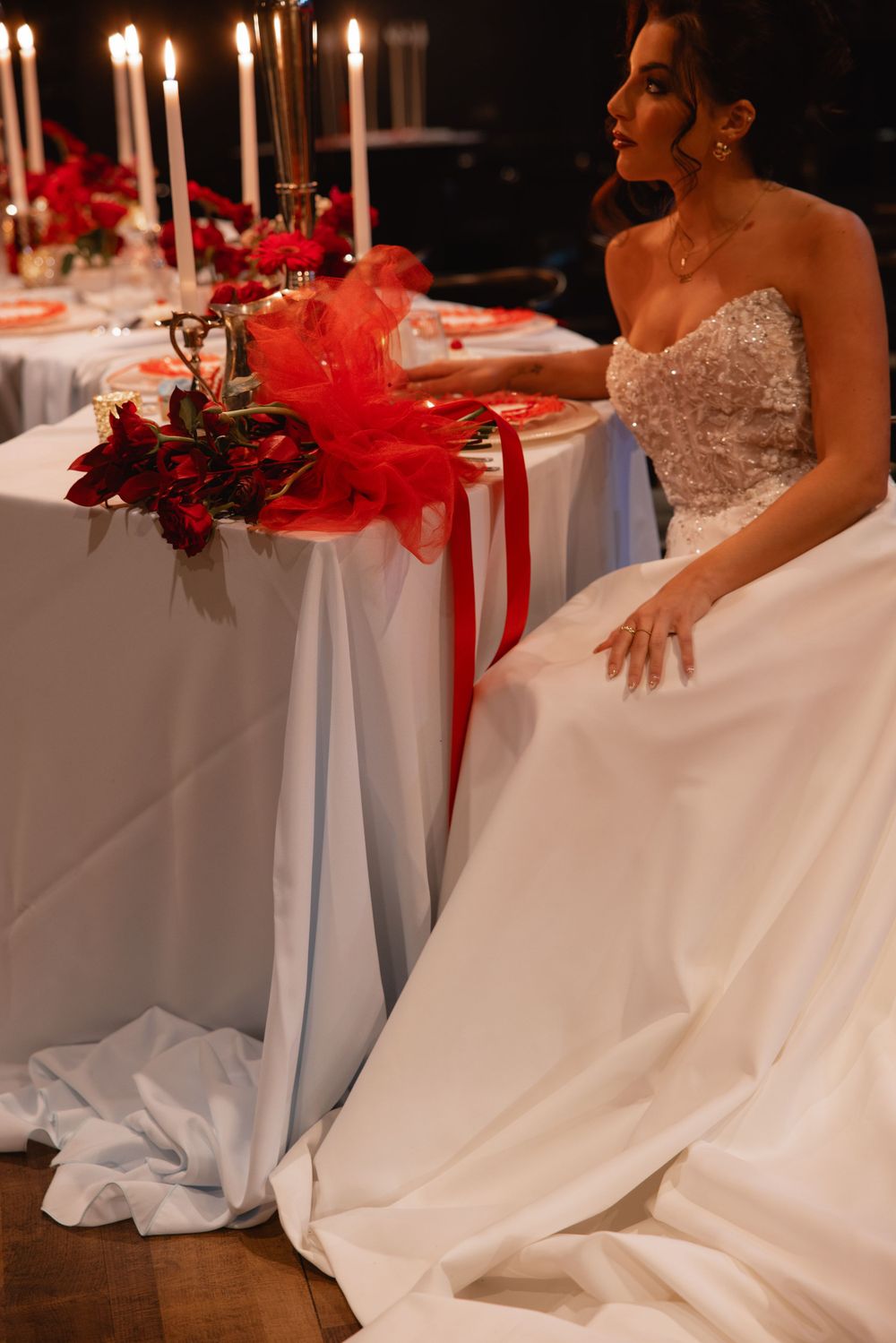 Elegant bride in a strapless beaded wedding gown standing beside a candlelit red and white reception table.