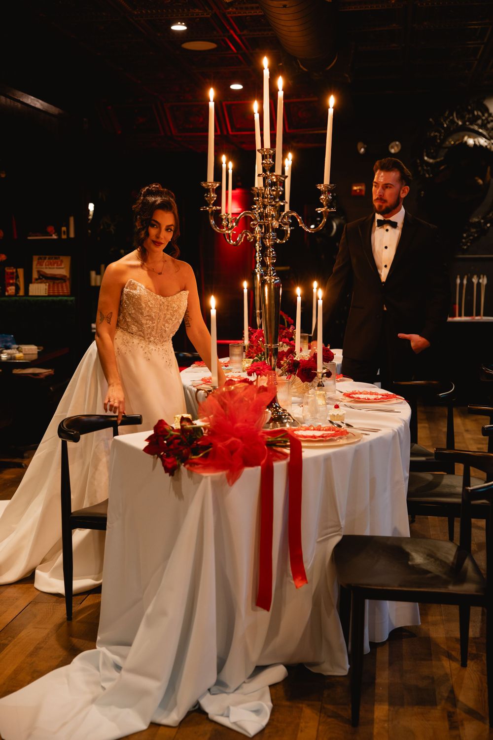Bride and groom in formal attire standing beside an elegant candlelit wedding table with red floral decor.