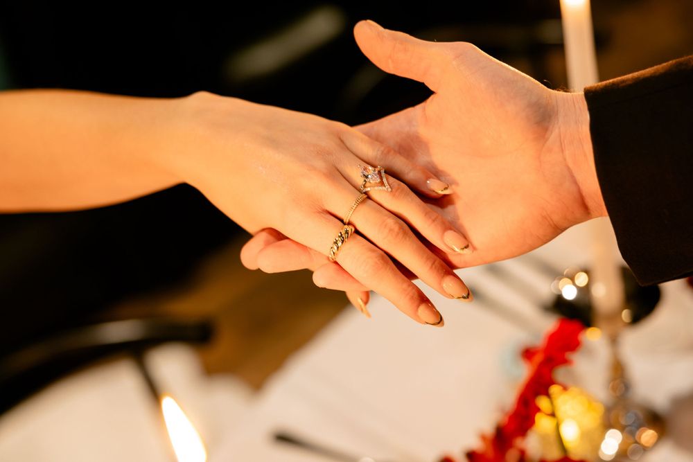 Close-up of bride and groom holding hands with a delicate gold engagement ring at a candlelit table.