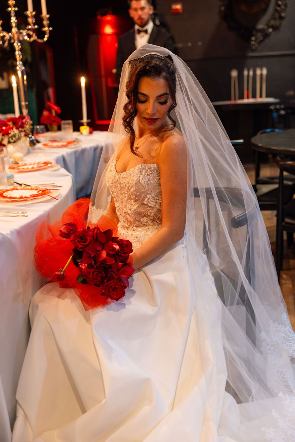 Beautiful bride in lace wedding dress with veil holding vibrant red rose bouquet at decorated reception table.